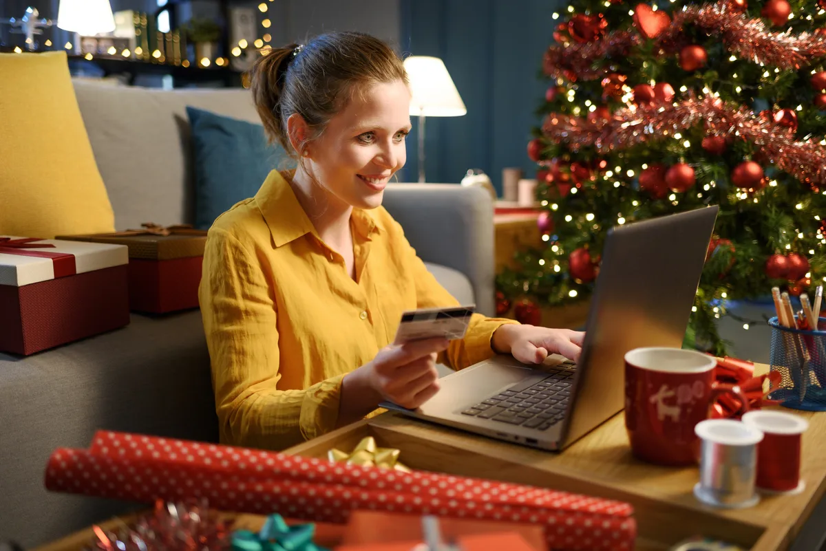 mujer en su casa decorada de navidad, frente al ordeandor con expresion feliz y la traketa dle banco en su mano por haber recibido el aguinaldo de navidad y declarar en hacienda