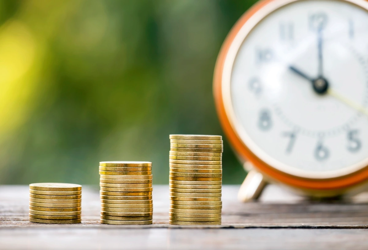 Monedas apiladas junto a un reloj, representando ahorro a largo plazo.